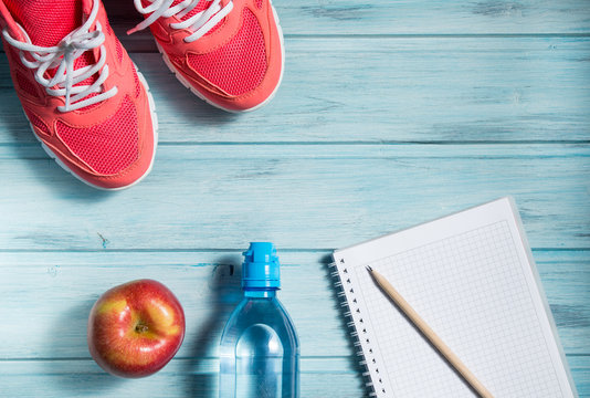 Fitness concept, pink sneakers, red apple, bottle of water and notebook with pencil on wooden background, top view