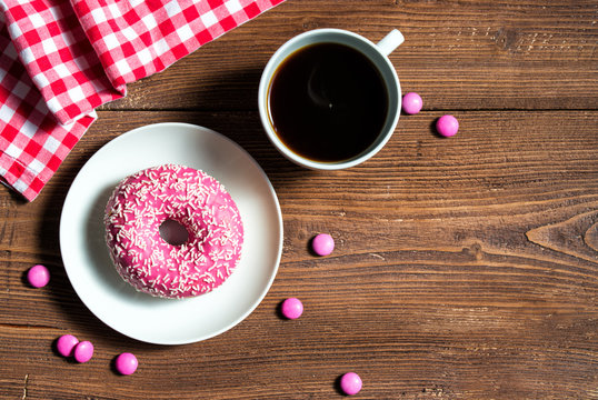 Pink donut on a plate with cup of coffee and pink candies, wooden background, top view