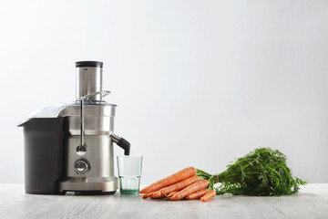 Side view metallic professional juicer with empty glass is prepared to make tasty juice for breakfast from fresh carrots lying on wooden table. Isolated on white background in cafe shop