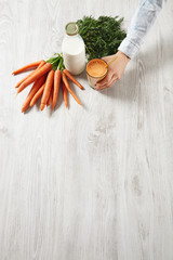Side top view on wooden table, farm carrot harvest lying near bottle and man hand holds glass filled with mix natural fresh juice and milk with golden drinking straw in it,for breakfast in cafe