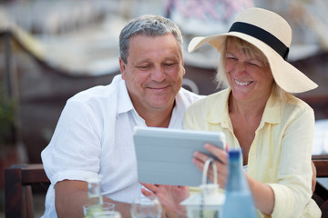 Smiling happy relaxed middle-aged couple using a tablet together on vacation as they sit at a table in a cafeteria enjoying refreshments