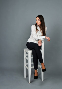 Beautiful young brunette girl sitting on a white chair. Pretty woman in white blouse