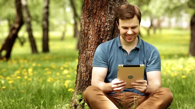 Pretty smiling hipster man using his tablet pc on a sunny day in park sitting on the grass with copyspace