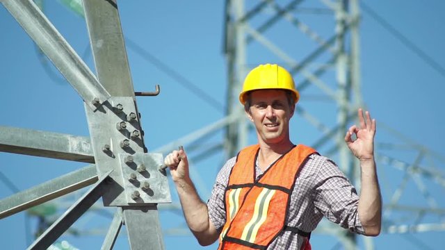 Closeup shot of a male technician in a safety vest and hard hat standing on the side of a high tension electrical tower visually inspects the situation and gives the viewer an OK gesture.