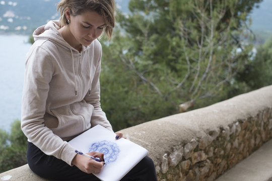 Young woman drawing a purple mandala pattern on a sketchpad with a pencil outdoors with the sea behind her