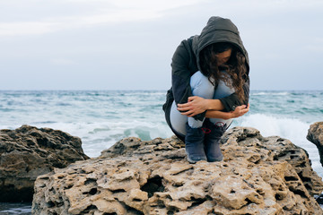 Girl in the green jacket with hood sits alone on a rocky on a co