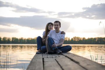 Smiling couple resting on the bridge on the shore of the lake at sunset
