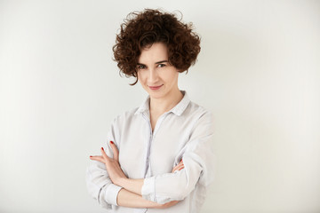 Isolated portrait of young happy confident female intrepreneur with short curly hair cut and brown eyes, posing with arms crossed against white copy space wall for your text or avdertising content