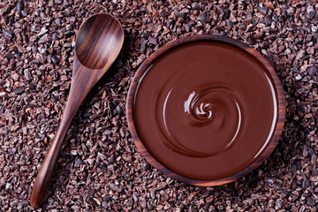 Bowl of melted chocolate and wooden spoon on a crushed raw cocoa beans, nibs background. Copy space...