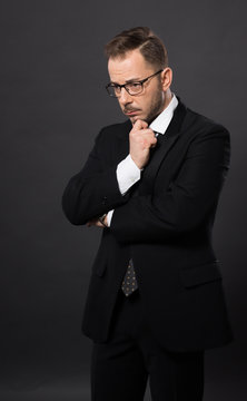 Portrait of thinkative businessman in glasses in studio. Handsome man in black suit looking down and dreaming about something.
