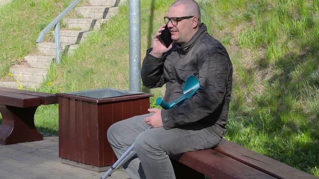 Disabled man with crutches sitting on bench and talking on smart phone