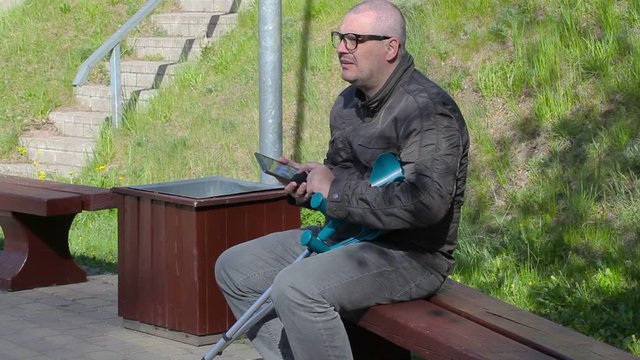 Disabled man with crutches sitting on bench and using tablet PC