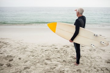 Senior man with surfboard shielding eyes at beach