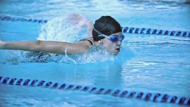 Side view of sportswoman in cap and goggles swimming butterfly stroke in pool in slow motion