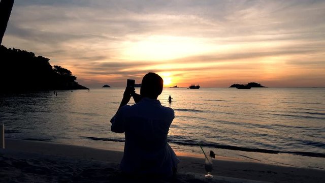 Male hand taking picture of sunset with cellphone on beach
