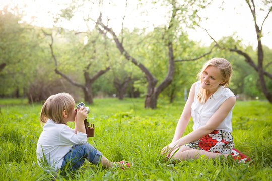 Adorable little kid boy sitting on the grass and taking picture of his young mother with the retro photo camera. Mother and son having photo session in the summer park. Outdoors.