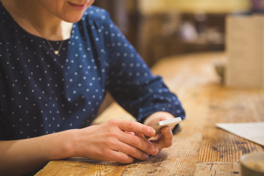 Close up photo of beautiful young woman sitting cozy at wooden table. Girl using  touch screen smartphone. Typing on the telephone.