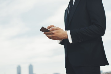 Professional employer using modern smartphone outside, close-up image of young successful businessman typing text message on his cellphone outdoors, blurred city in the background