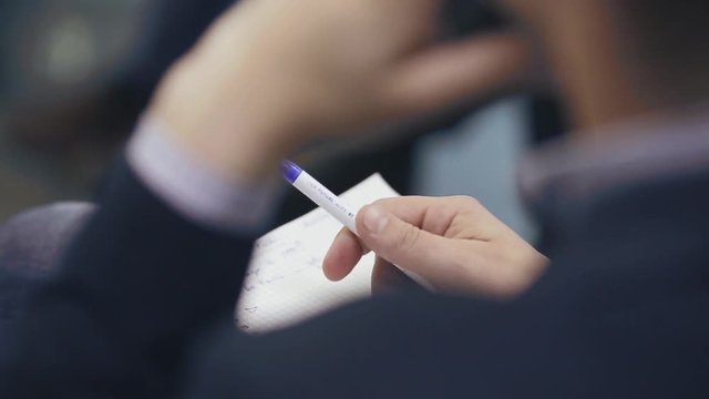 Close up, over the shoulder shot of a business man holding a pen, thinking and making notes in notebook during business meeting or conference or training. 
