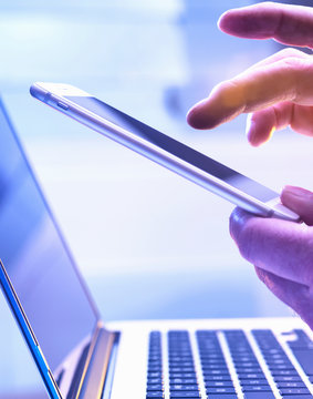 Man operating a touch screen mobile phone with a laptop in the background.