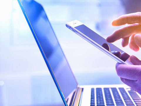 Man operating a touch screen mobile phone with a laptop in the background.