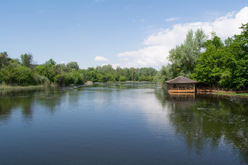 Fototapeta na wymiar River bank: spring landscape. Blue sky, green trees and small wooden summerhouse on the water