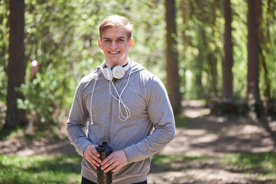 Portrait of a smiling athlete with headphones around his neck holding the shaker in hand on a forest background