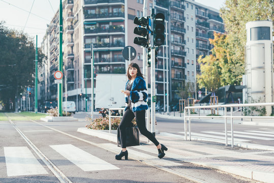 Young handsome eastern woman walking outdoor in the city crossing pedestrian crossing, holding a smart phone and a bag, overlooking - technology, social network, business concept