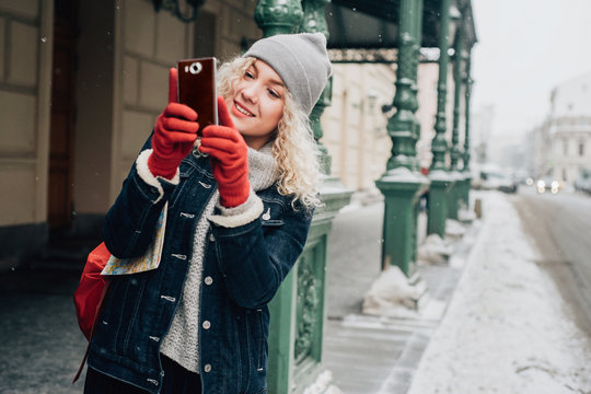 Young blond curly female tourist in warm clothes and red gloves photographs or taking a selfie on city street, winter, snow