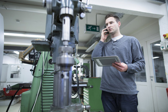 Young male technician chatting on smartphone whilst using digital tablet in workshop