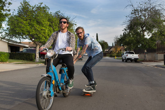 Male skateboarder skitching on friends moped on suburban road