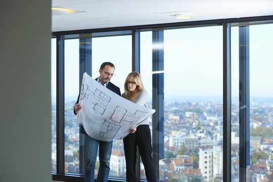 Two architects looking at plans in front of office window with Brussels cityscape, Belgium
