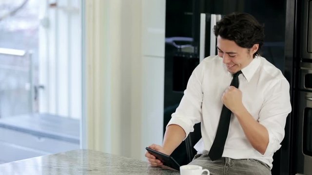 A Handsome Businessman In His 20s Wearing A White Shirt And Tie, Sitting Down In His Kitchen Working From Home. Successful, Satisfied And Happy.