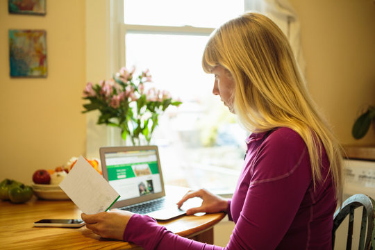 Woman working in kitchen table using laptop and  reading instruction booklet