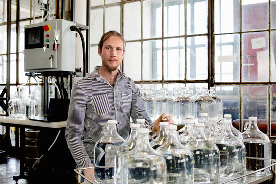 Portrait of young male vodka distiller and demijohns in distillery workshop
