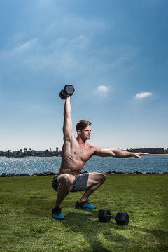 Mid adult man crouching and lifting dumb bells at coast