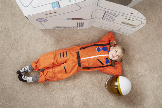 Young girl playing, wearing astronaut outfit, lying next to cardboard spaceship, elevated view