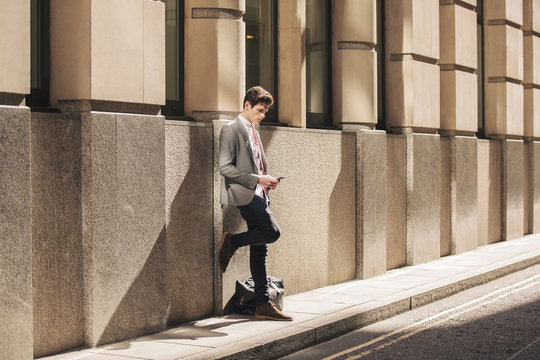 Young city businessman leaning against office building reading smartphone text