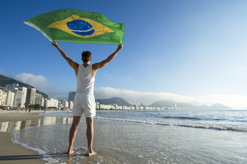 Athlete in white outfit standing with Brazil flag waving in the wind on the shore of Copacabana Beach, Rio de Janeiro, Brazil
