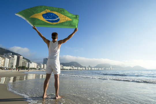 Athlete in white outfit standing with Brazil flag waving in the wind on the shore of Copacabana Beach, Rio de Janeiro, Brazil
