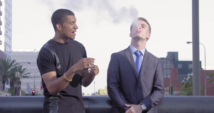 Two young men, African American and his Caucasian friend in a suit talk against backdrop of Downtown Los Angeles. Businessman smokes e-cigarette.