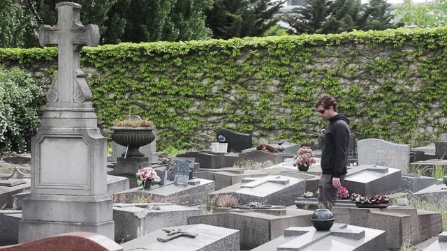 Man Visiting Tomb In Cemetery. Young man arrives in a cemetery and visits a tomb next a big holy cross