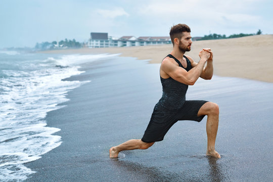 Fitness Exercise. Sporty Handsome Male Stretching Legs Before Run. Fit Athletic Man With Muscular Body Exercising On Sand, Training During Outdoor Workout At Beach. Sports, Healthy Lifestyle Concept 