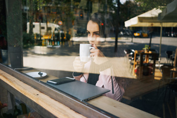 attractive girl enjoying a morning coffee while sitting at a table in a cozy cafe. Portrait of a young girl through the panoramic window in the coffee shop. smartphone and laptop on the table.