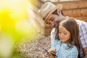 Girl on fathers lap using smartphone in community garden