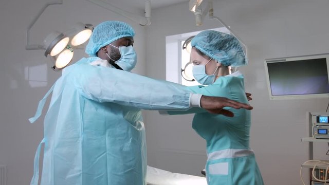 Female caucasian nurse don sterile disposable surgical gown on male african surgeon.