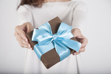 brown gift box with blue ribbon in woman blue jeans cream jersey hands over white background

