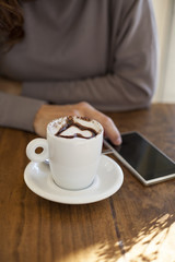 woman hand touch phone next to white cup cappuccino coffee with chocolate heart on light brown wooden table

