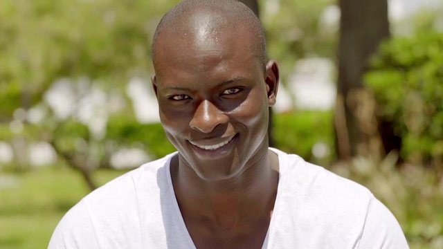 Smiling handsome bald black man in the park while wearing white shirt near hedge and trees