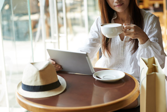 Cropped image of young woman with tablet enjoying cup of cappuccino in cafe